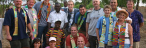 A group of people pose outdoors, many wearing colorful patterned sashes, with trees in the background.