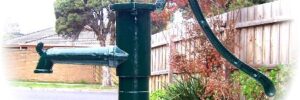 A black metal hand water pump is installed in a garden near a wooden fence and some trees.