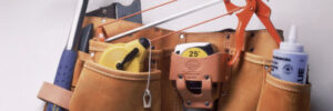 Leather tool belt containing a hammer, saw, tape measure, glue bottle, and other small tools on a white background.