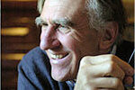 An older man with gray hair and a sweater sits indoors, smiling and holding something in his right hand.
