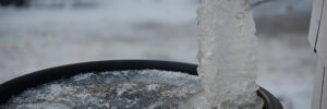 A frozen downspout with solid ice extends into a barrel partially covered with ice, outdoors in winter.