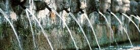 Stone fountain with multiple sculpted heads spouting streams of water into a shallow basin below.