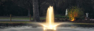 A lit water fountain sprays up from the center of a pond, surrounded by trees and a statue in the background.