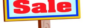 A "For Sale" sign with red text on a white and blue board, placed on a wooden post in green grass.