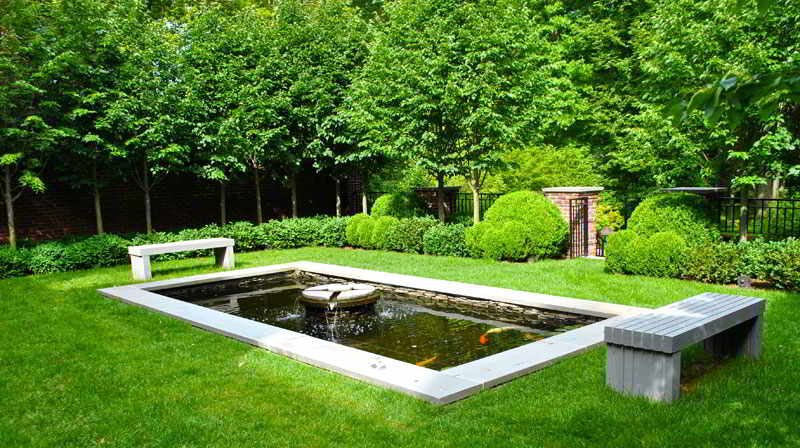 Rectangular pond with a small fountain in a landscaped garden, bordered by two benches and surrounded by green trees and shrubs.