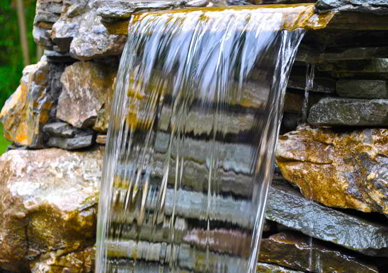 Water flows over a stone ledge in a small waterfall, with stacked rocks visible behind the falling water.