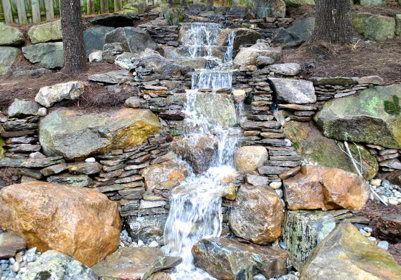 A small artificial waterfall flows over stacked rocks and stones in a landscaped garden setting.