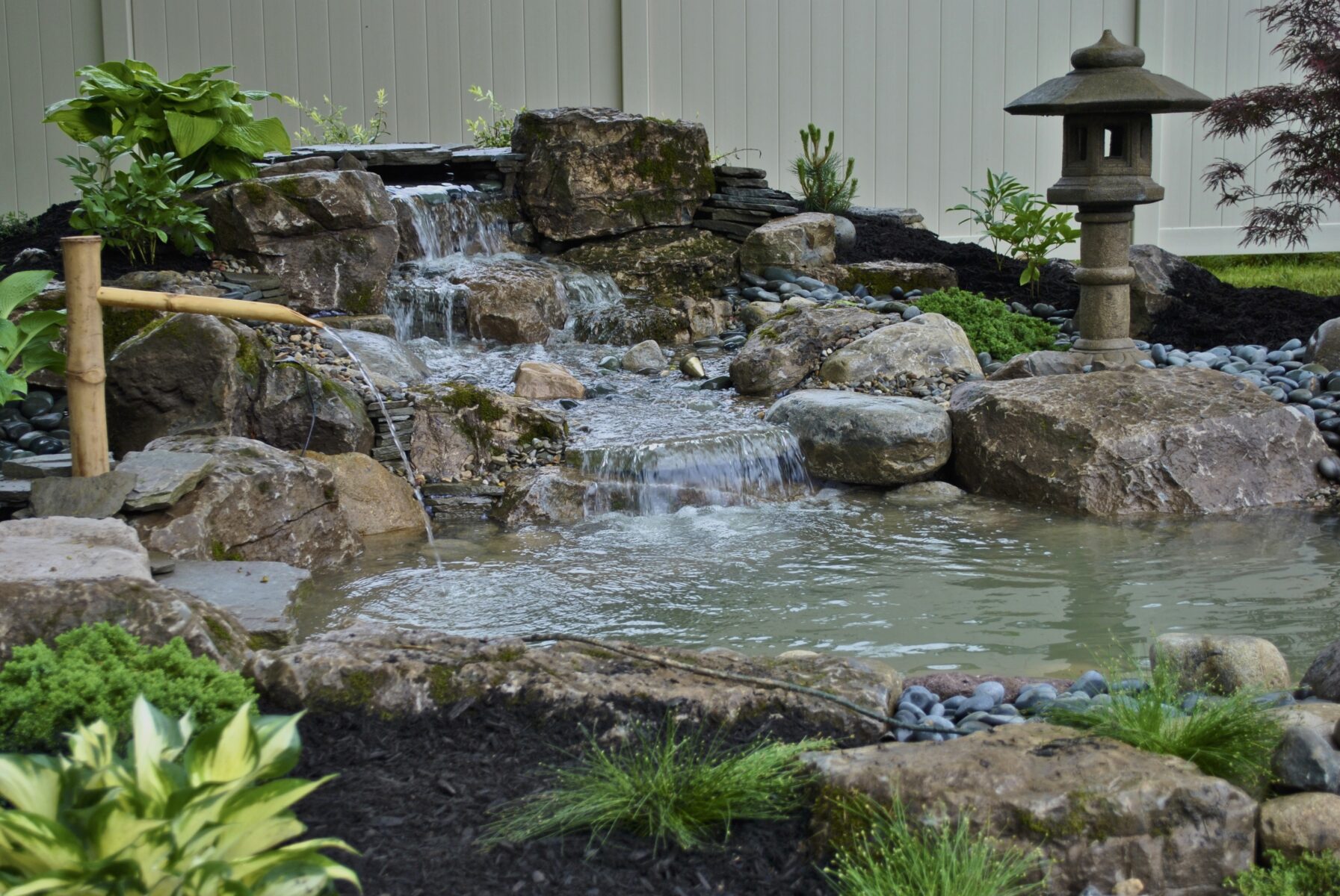 Small backyard pond with a rock waterfall, bamboo fountain, and stone lantern, surrounded by plants and mulch.