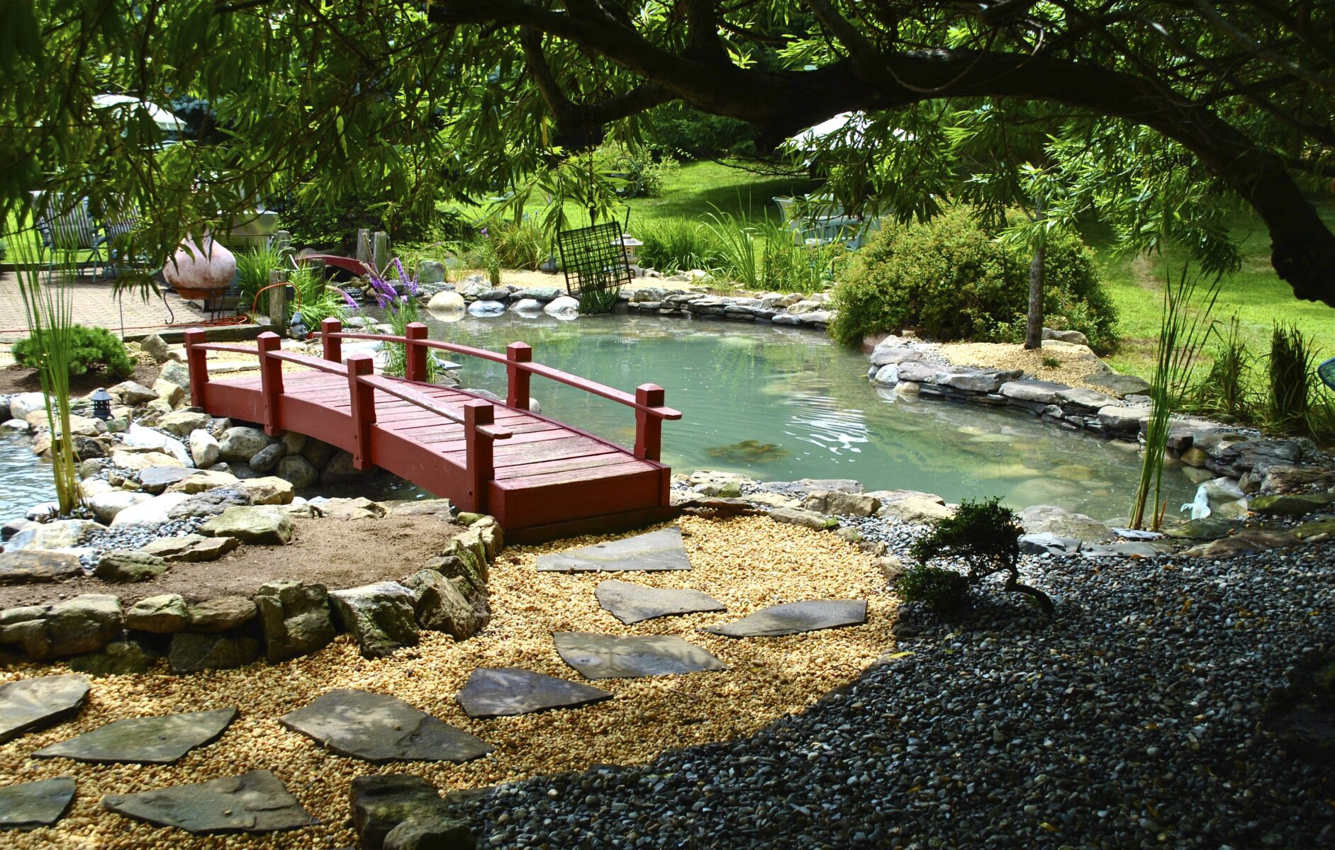 A small wooden bridge crosses over a pond in a landscaped garden with rocks, gravel paths, and surrounding greenery.