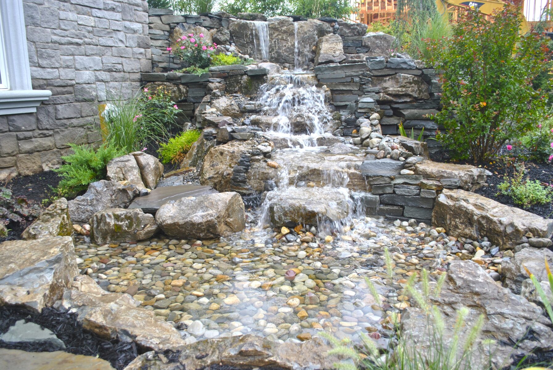 Water flows down a tiered stone waterfall into a shallow pond surrounded by rocks and garden plants.