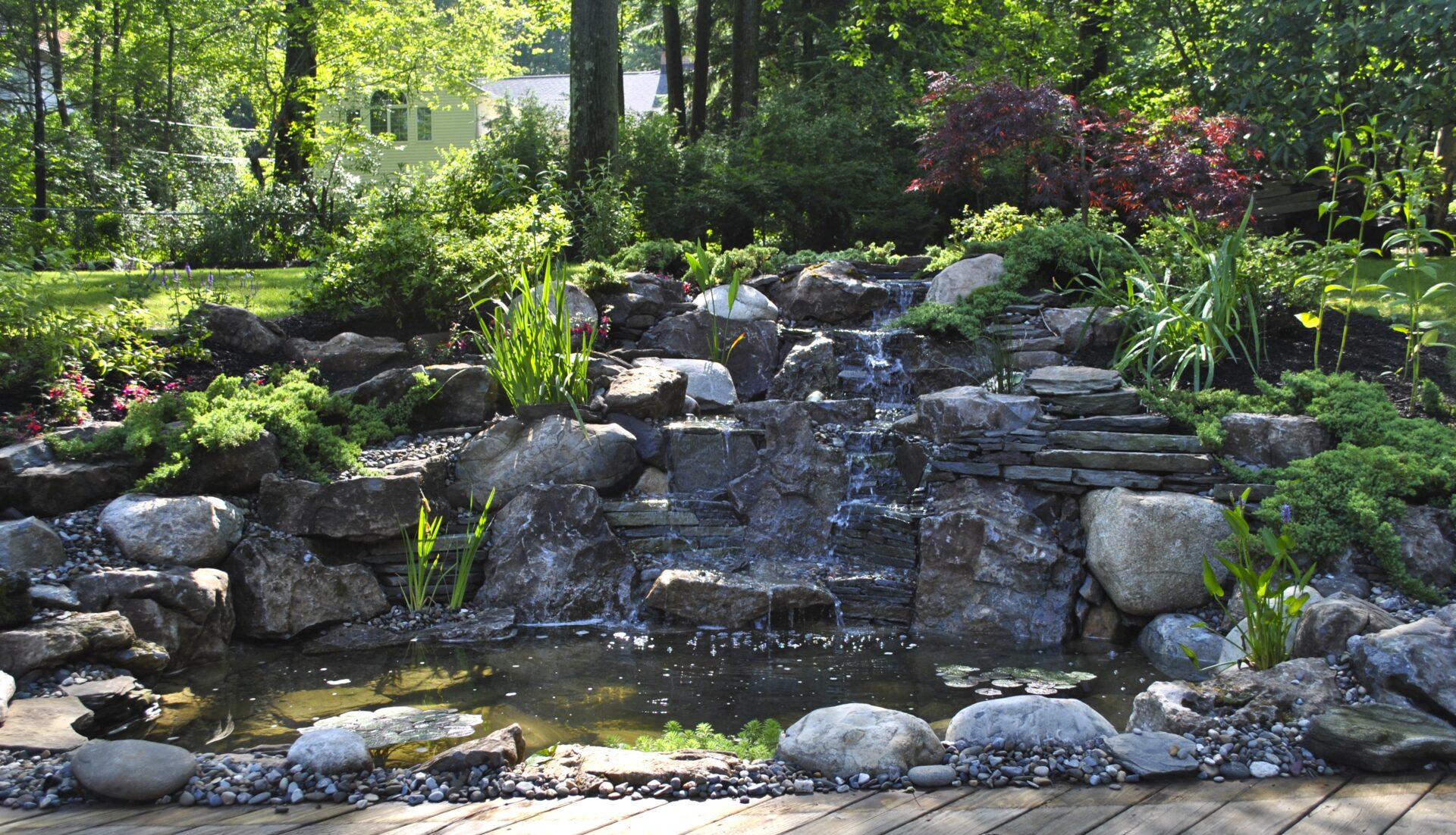 A backyard garden with a stone waterfall, pond, lush green plants, and sunlight filtering through the trees.