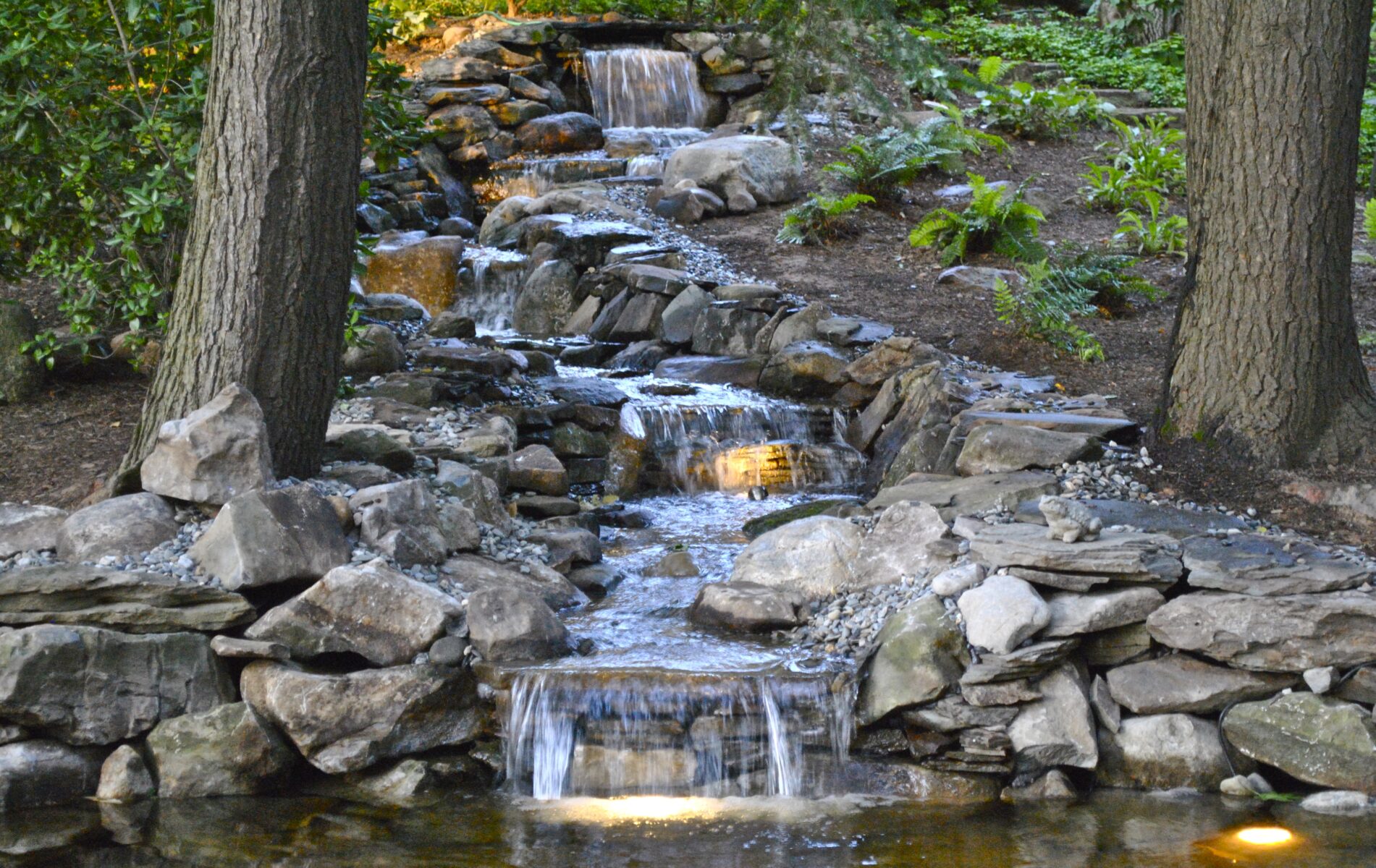 A small, tiered waterfall flows over rocks between trees, surrounded by greenery, with lights illuminating the water.