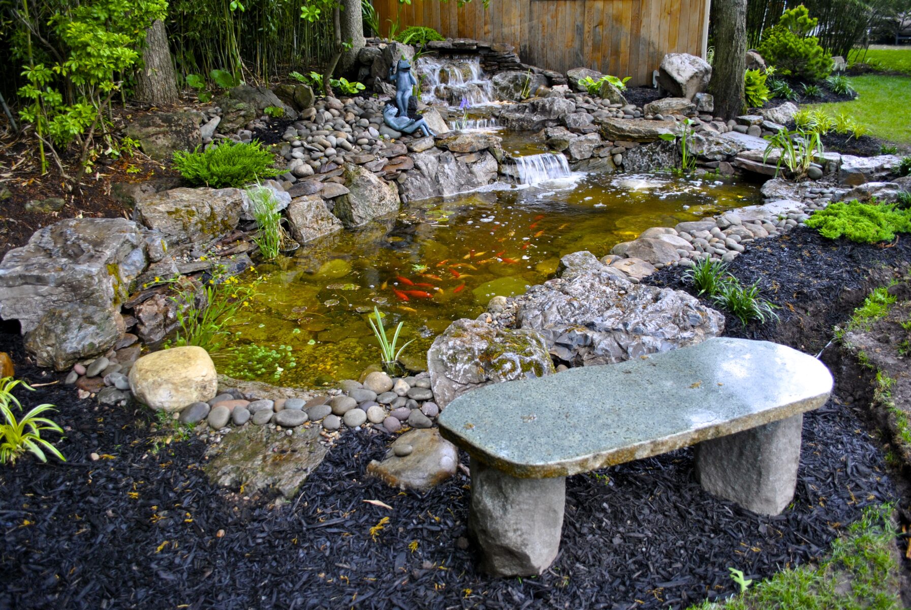 Stone bench facing a small koi pond with a waterfall, surrounded by rocks, plants, and a wooden fence in a garden.