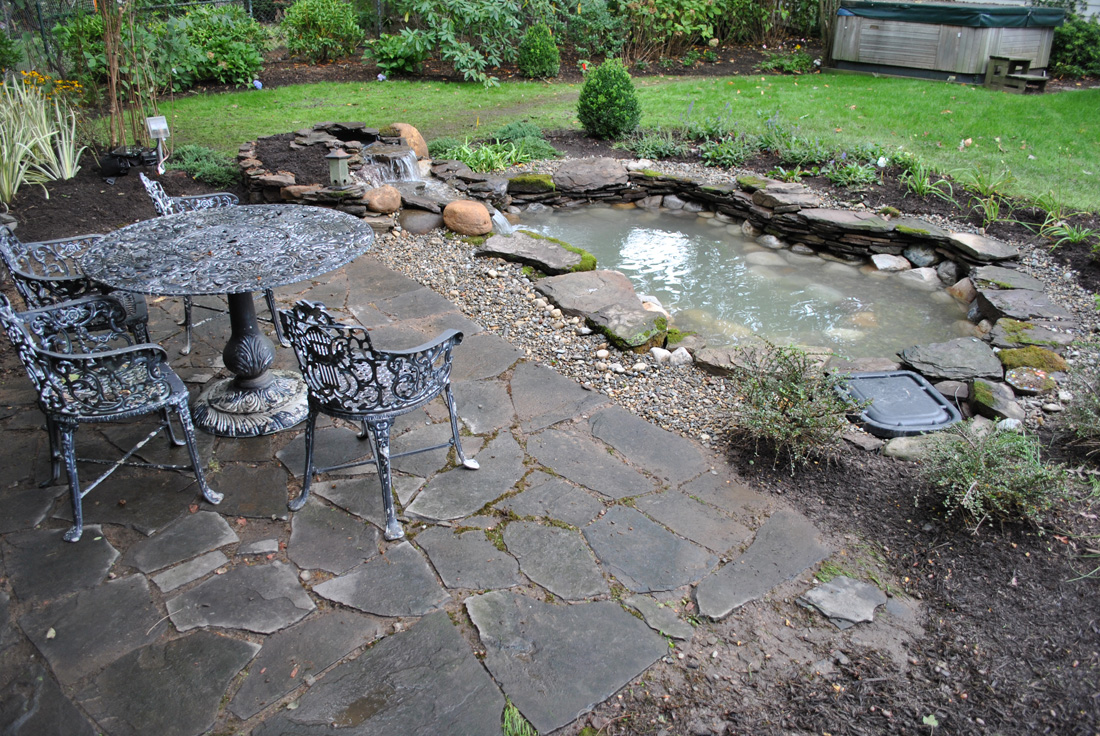 A stone patio with a metal table and chairs overlooks a small backyard pond bordered by rocks and plants.