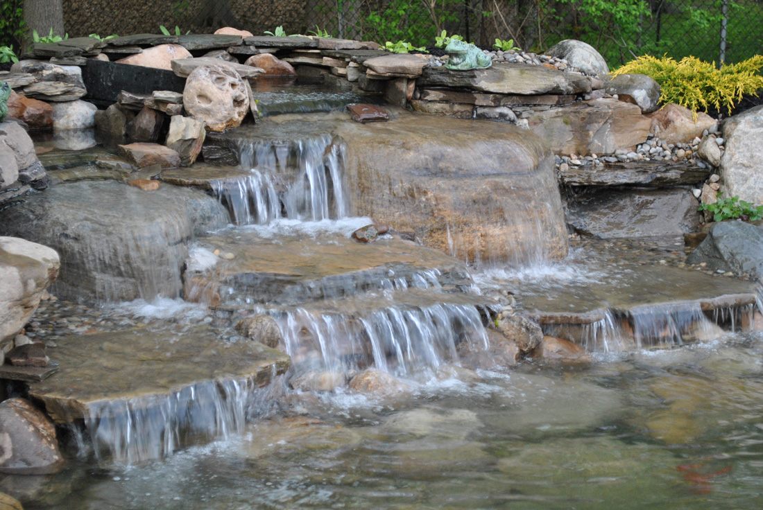 Water cascades over stone steps in a small garden waterfall surrounded by rocks and greenery.