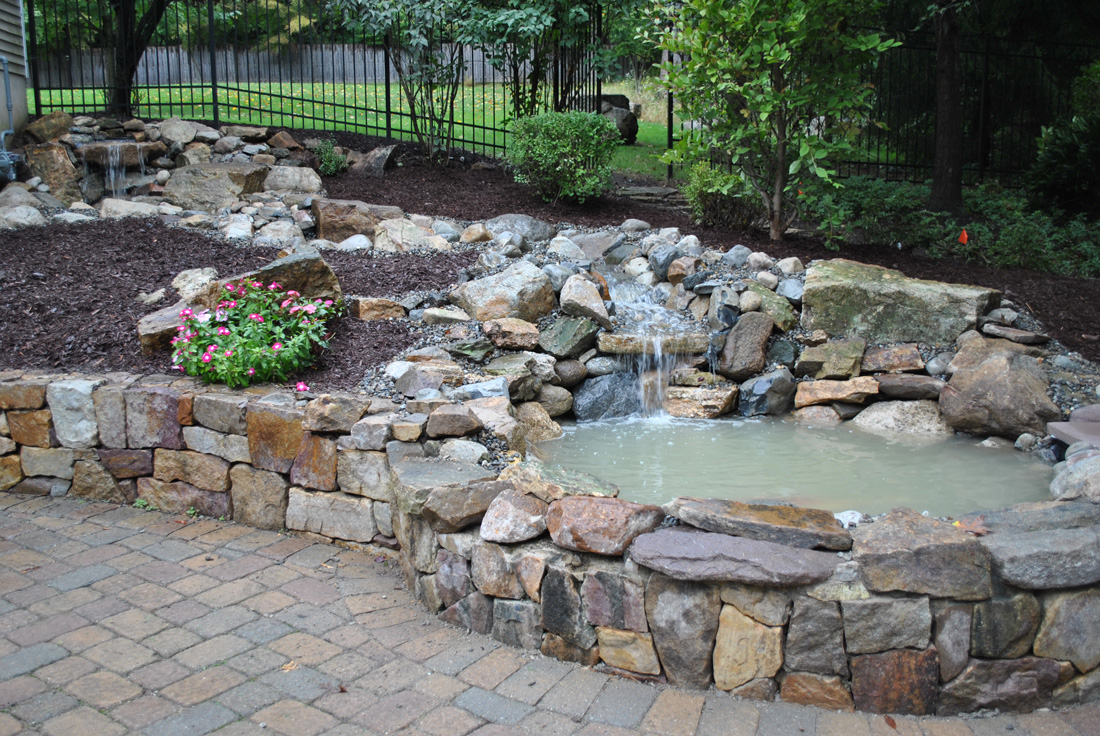 Small backyard pond with waterfall feature, surrounded by rocks, mulch, flowers, and stone paver patio.