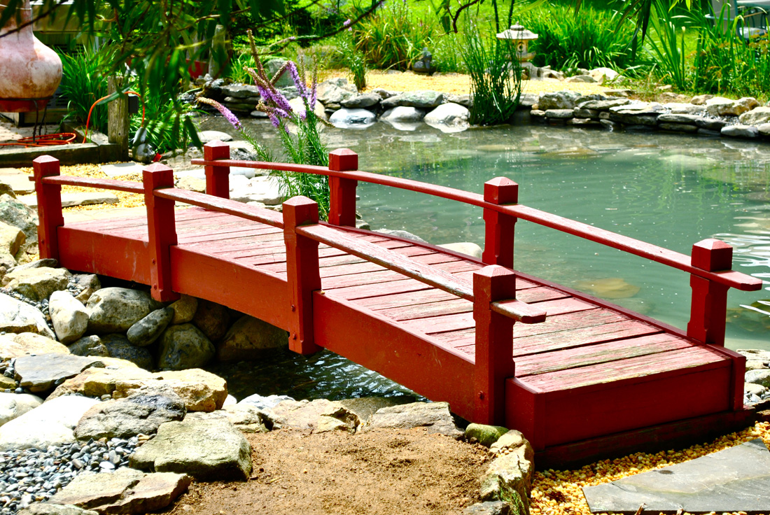 A small red wooden bridge crosses over a pond surrounded by rocks and greenery in a garden.