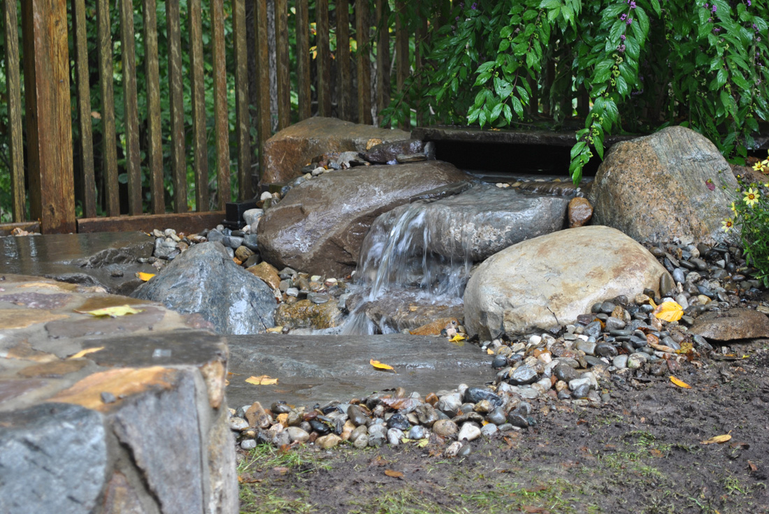 Small waterfall flows over rocks in a landscaped garden with a wooden fence and leafy green plants in the background.