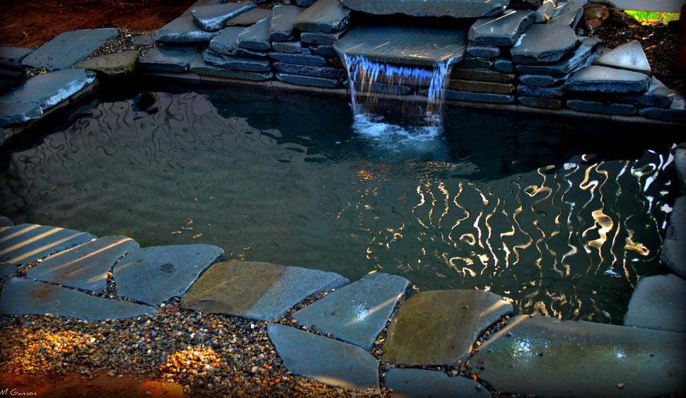 A small stone-lined pond with a short waterfall, surrounded by rocks and gravel, reflecting light on the water’s surface.