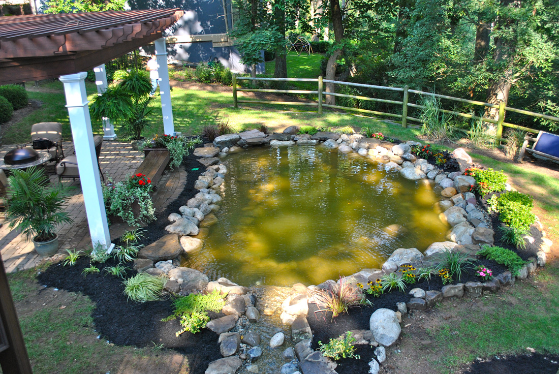 Backyard with a stone-edged pond, surrounding plants, patio seating, and wooden fence in a wooded area.