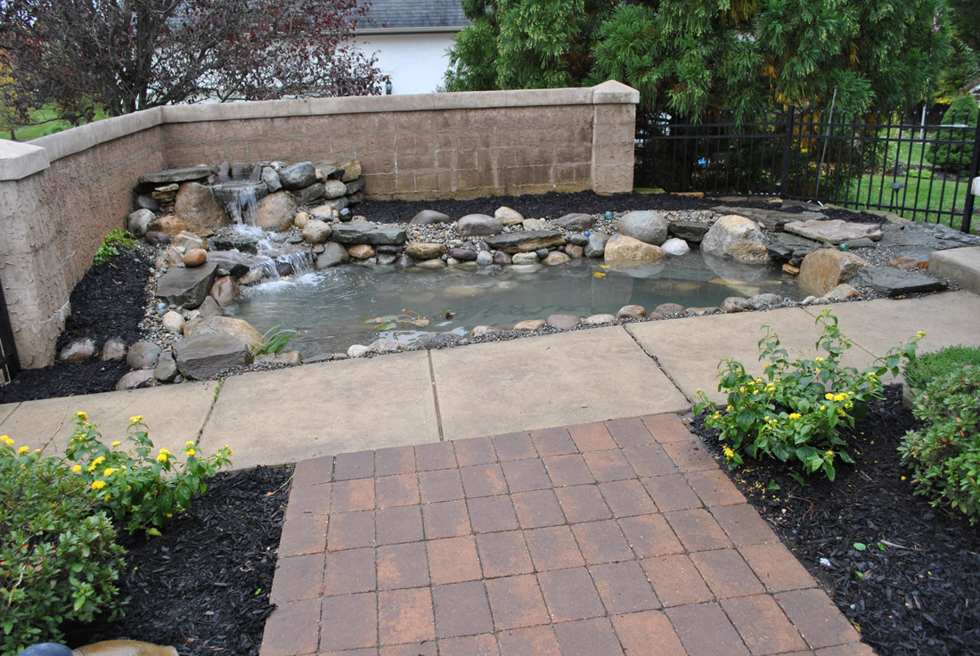 A small backyard pond with a stone waterfall feature, surrounded by a paved walkway and landscaped plants.