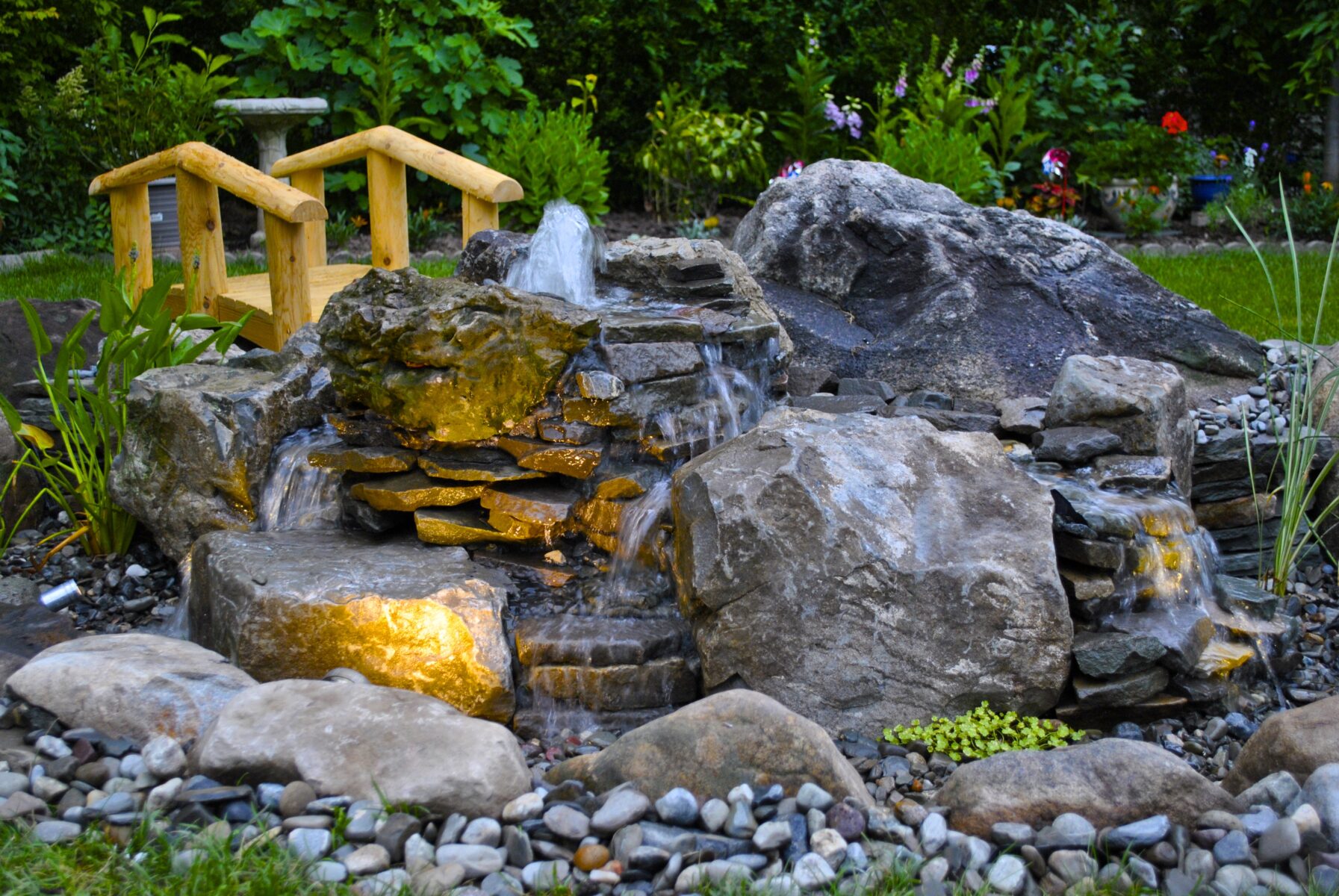 Small backyard pond with waterfall feature, rocks, small wooden bridge, and garden plants in the background.