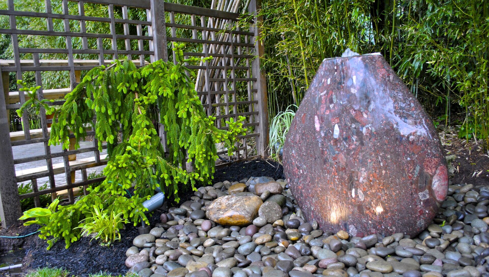 Large polished stone water feature surrounded by smooth rocks, greenery, and a wooden lattice fence in a garden setting.