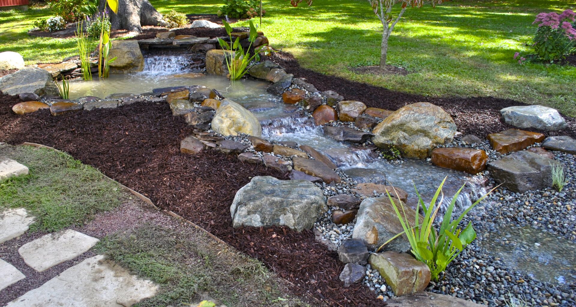 A small artificial stream with rocks and plants flows through a landscaped garden with mulch and grass.