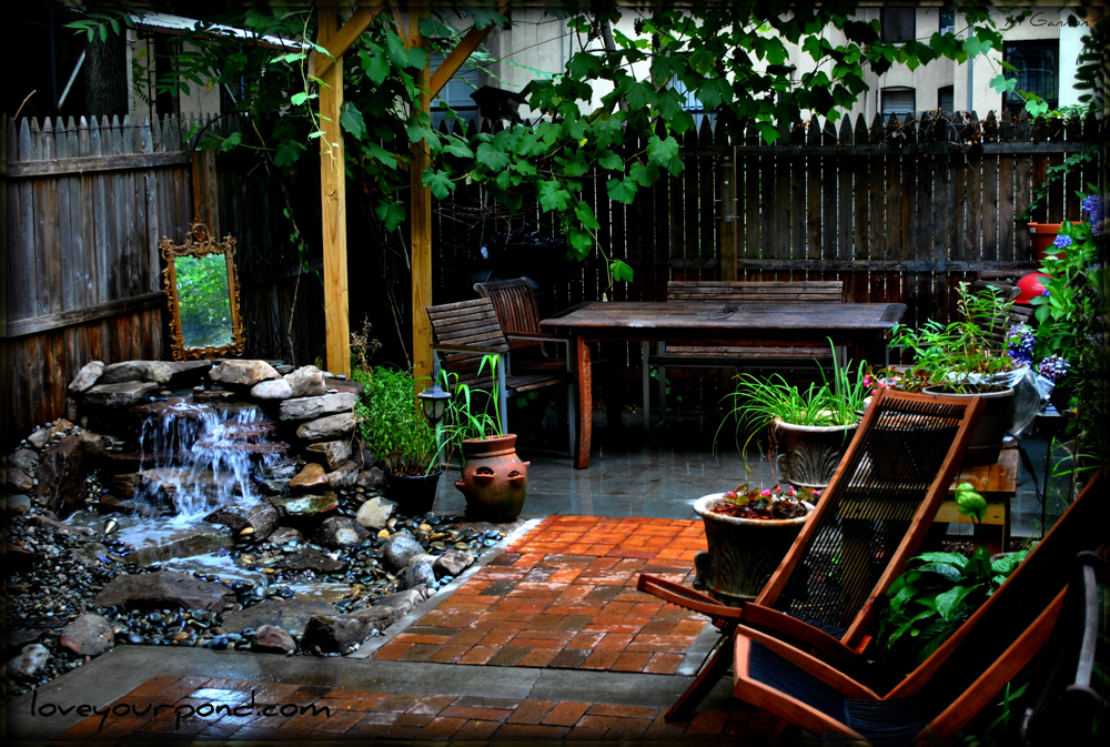 A backyard patio with wooden furniture, a small waterfall feature, potted plants, and a wooden privacy fence.