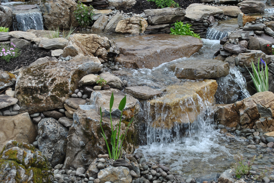 Small waterfall flowing over rocks surrounded by plants and pebbles in a landscaped garden setting.