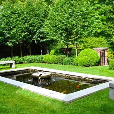 Rectangular pond with a small fountain in a landscaped garden, bordered by two benches and surrounded by green trees and shrubs.