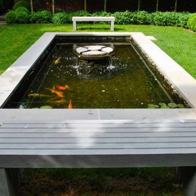 Rectangular koi pond with concrete border and benches, featuring water lilies and an art piece in the center.