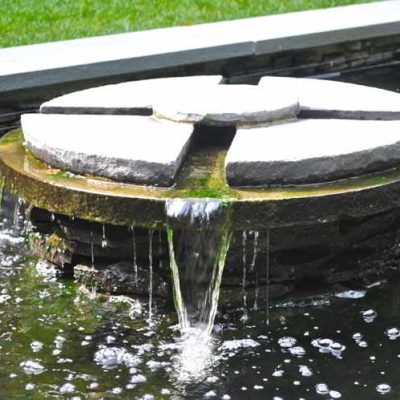 Round stone water fountain with segmented top, water flowing over edges into a shallow pool below, grass in background.