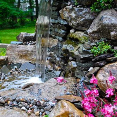 A small waterfall flows over stacked rocks into a pond, with pink flowers and green plants in the foreground.