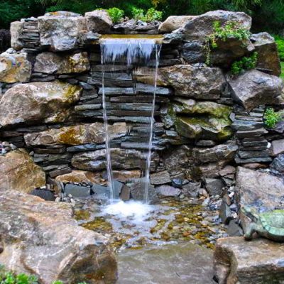 Small artificial waterfall flows over stacked rocks into a shallow pond, surrounded by greenery and a turtle statue.