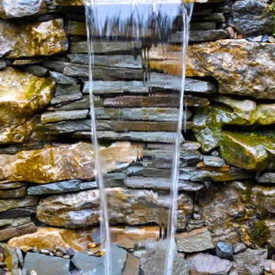 Water flows in a thin stream over stacked rocks, forming a small waterfall in a stone garden wall.