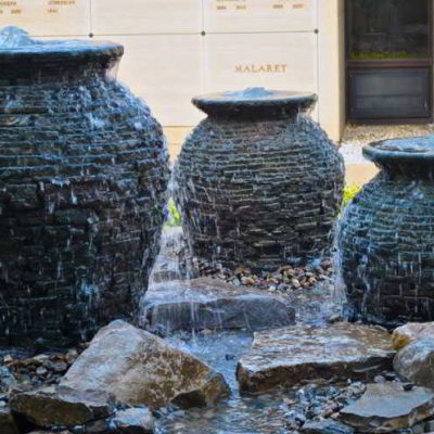 Three large stone urn fountains with water cascading down their sides, surrounded by rocks and a stone wall background.