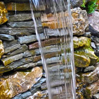 Water flows down a stone wall, forming a small waterfall with rocks and greenery surrounding the feature.