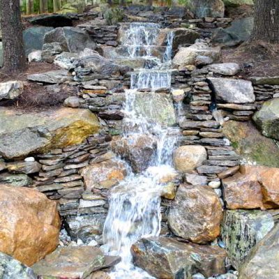 A small artificial waterfall flows over rocks and stone slabs in a landscaped garden area.