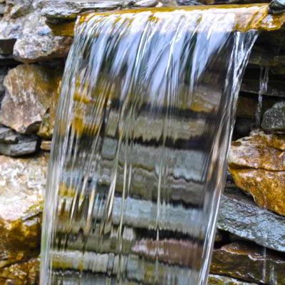 Water flows over a stone ledge in a small waterfall, with stacked rocks visible behind the falling water.
