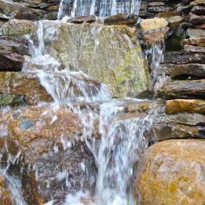 Water cascades down layered rocks, forming a small waterfall with clear, flowing water surrounded by stones.