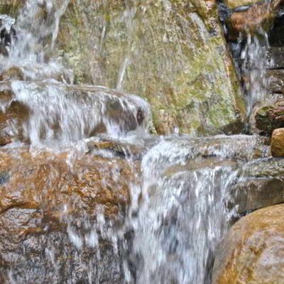 Water flows over stacked rocks in a small, tiered waterfall, creating white splashes and wet stones.