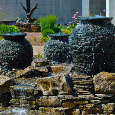 Three stone urn fountains with water flowing over them sit on a stone base in a landscaped garden.