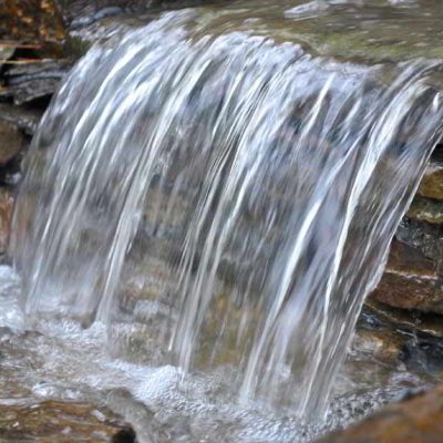A small waterfall flows over layered rocks, creating ripples and splashes in the clear water below.