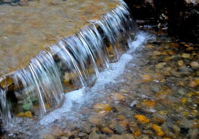 Clear water gently cascades over a flat rock ledge into a shallow pool with visible stones beneath the surface.
