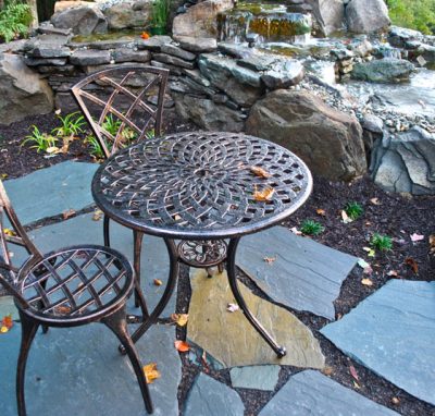Metal patio table and two chairs on a stone terrace beside rocks and a small pond with scattered leaves.