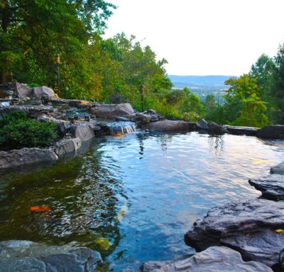Natural stone pond with small waterfalls, surrounded by trees and overlooking a distant valley under a clear sky.