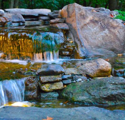 Rock waterfall with small cascades and underwater lights flowing into a shallow pond, surrounded by large stones and greenery.
