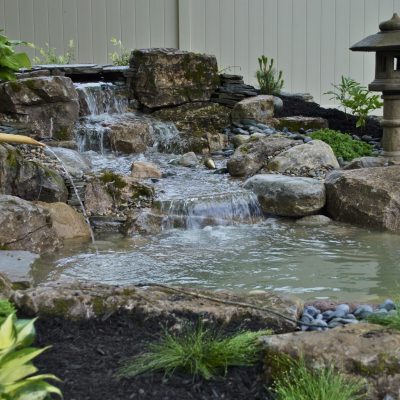 Small backyard pond with a rock waterfall, bamboo fountain, and stone lantern, surrounded by plants and mulch.