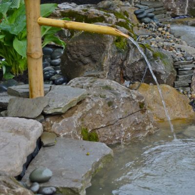 A bamboo water spout pours water into a small pond surrounded by large rocks and green plants.